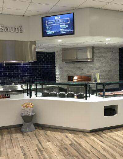 A modern cafeteria station with a sauté and pizza section, featuring a pizza oven and digital menu displays.