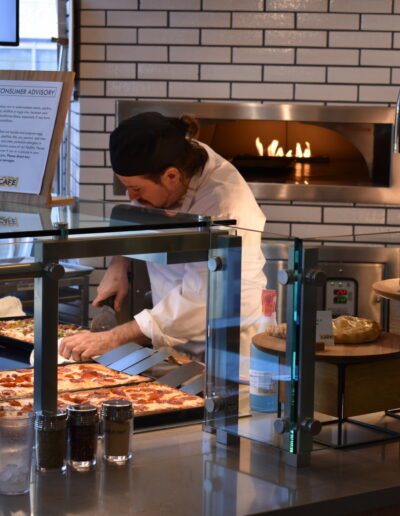 A chef preparing food behind a glass partition in a pizza kitchen with ovens.