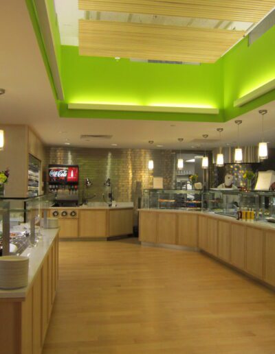 Modern cafeteria interior with food display, seating area, and bright green accent lighting.