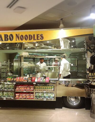 A noodle station named "tabo noodles" with a chef preparing food behind a glass counter, flanked by displays of packaged foods.